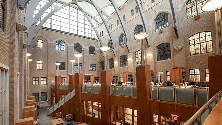 Interior of brick space with four floors and steel arches and large windows and skylights. Wooden half-columns support mezzanine level with green shaded panels in railing