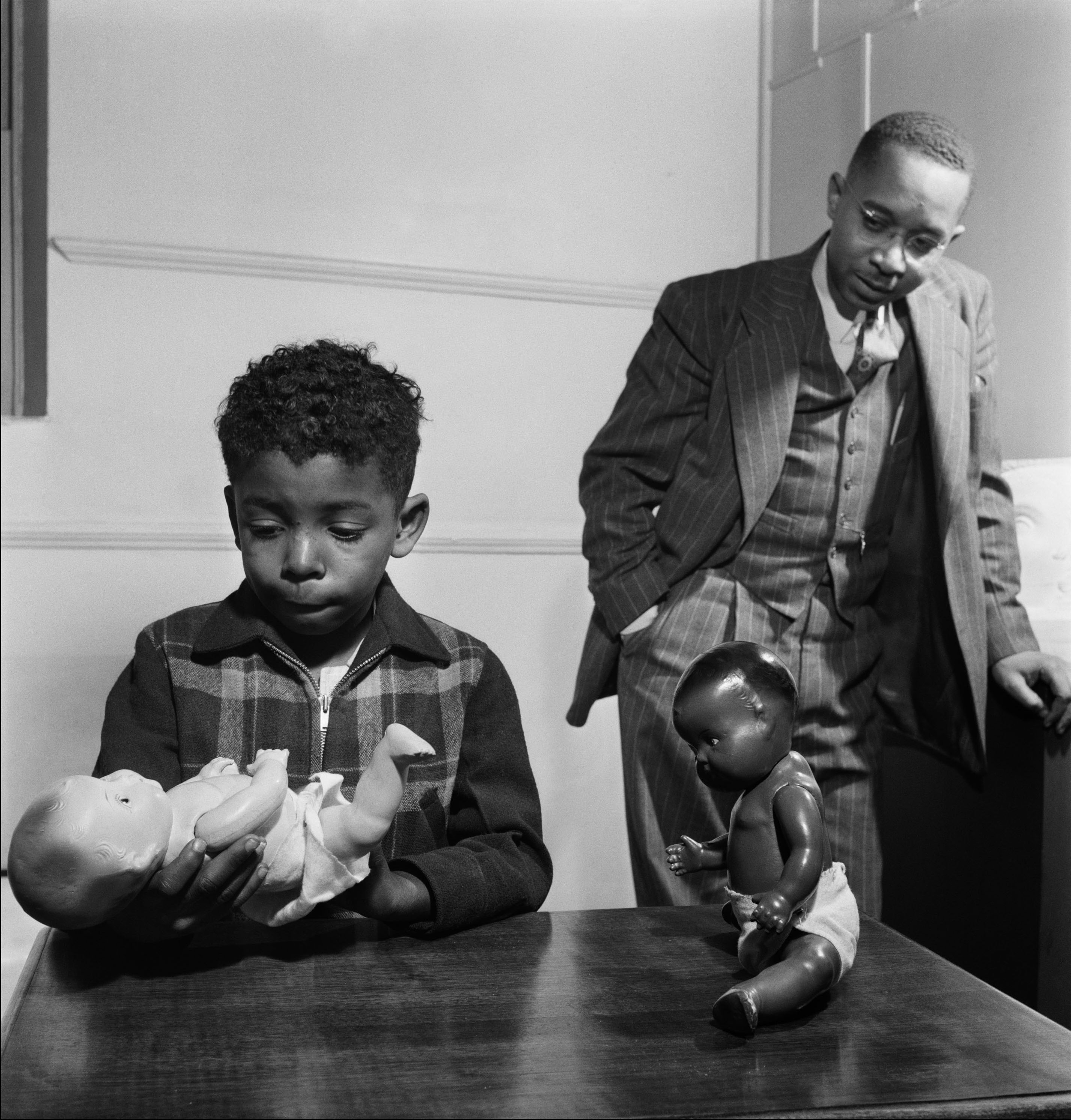 Black boy holds and looks at white baby doll with a Black baby doll sitting nearby on the table and a man in a suit stands in the background observing.