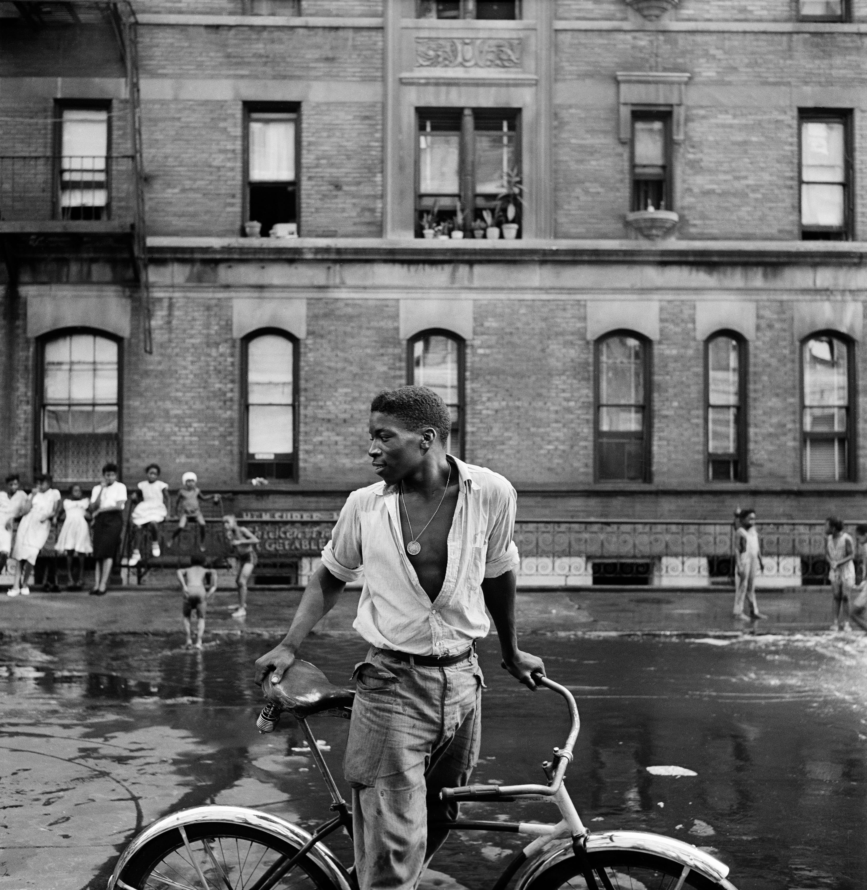 Young black man in white shirt, medallion and cargo pants stands astride a bike looking over his shoulder and smiling. In. background are brick apartment building, women leaning against a fence, and children playing in puddles.