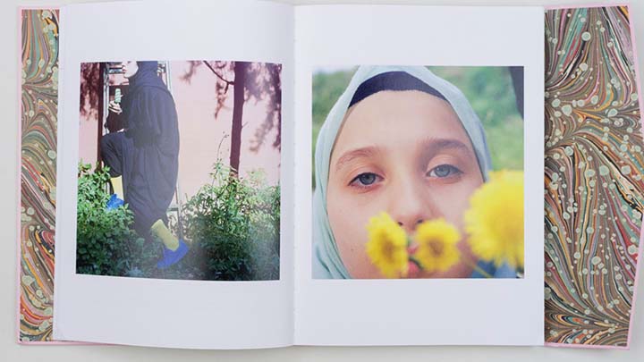 Inside book pages showing marbled papers inside front and back covers. Photo on left page shows woman in black Turkish dress with green socks and blue shoes stepping up a ladder in a garden with greenery around her feet; photo at right shows face of young girl looking at camera with three yellow flowers in the foreground in front of her mouth.