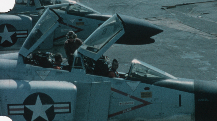 Aircraft on ground, with large white star in blue circle on side, with both hatches swung open and two pilots inside. A ground technician wearing goggles is talking to the pilot in the rear cockpit.