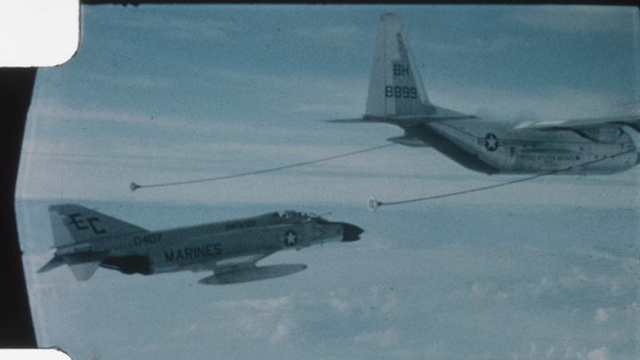 Two Marine jets in flight, with two long refueling cables from one extending to the smaller jet flying alongside