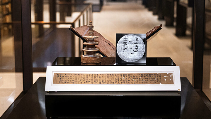 Close-up view of a glass display glass that shows a small yellowed horizontal scroll with Japanese characters in black ink. The scroll is in a white frame. Sitting behind the frame on a raised surface is a miniature wooden pagoda and a photograph showing the underside of the pagoda and Japanese characters there that spell the pagoda maker's name