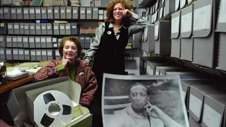 Three people in storage room with shelves of gray boxes. A woman with red hair stands; older woman with red and green jacket sits. In foreground is black and white photo of Aaron Copland. All three hold a finger to their jawline.