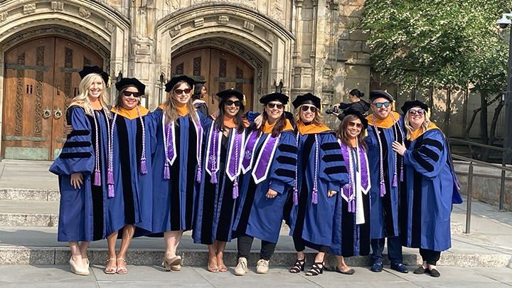 Nine graduates wearing dark blue robes with three black stripes along each arm, gold fabric at neckline and black caps pose in front of stone library entrance. All but the blonde woman at far left wear dark sunglasses.