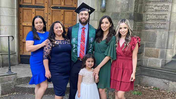 male with graduation cap at center with two women in blue dresses to his right, two younger women to left, one in green dress, one in red, and a little girl in front of him with a white dress