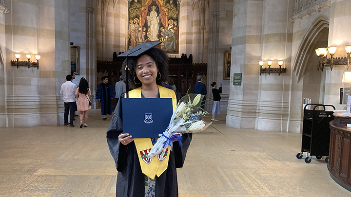 Female graduate stands inside nave with painting of Alma Mater behind her. Six people walking in distance have back to camera. She wears a cap and gold stole and holds her blue diploma folder and a bouquet of white flowers tied with blue ribbon.
