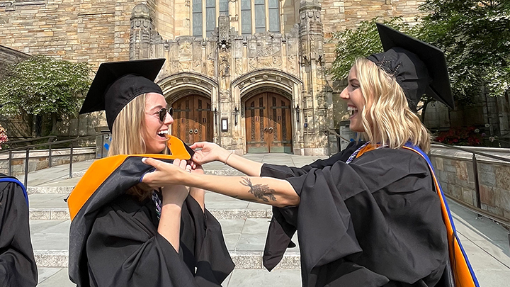 Two blonde women in front of library entrance, laughing. Woman on right is adjusting the gold stole of woman on left, who wears cap and sunglasses. Womon on right also wears cap and has tattoo on forearm.