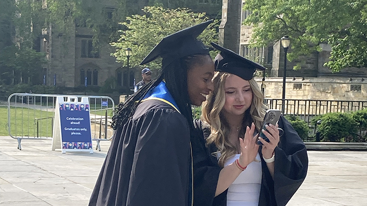 two female graduates looking at iphone screen, woman on right in white dress with blonde hair is touching the screen and has long fingernails with white polish