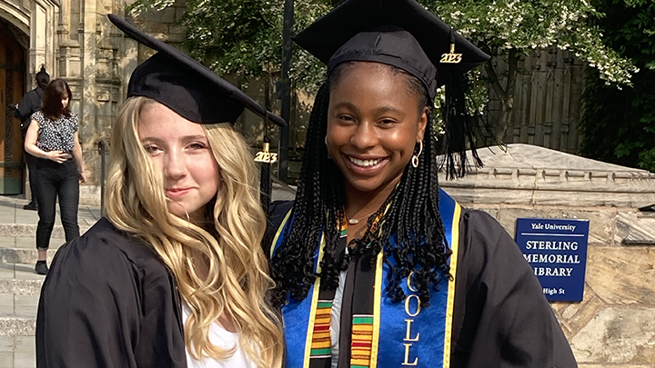 A woman with long blonde hair and tilted graduation cap stands next to woman with long black braided hair wearing a cap and a bright blue stole over her black gown. The background sign reads "Yale University Sterling Memorial Library, High St."