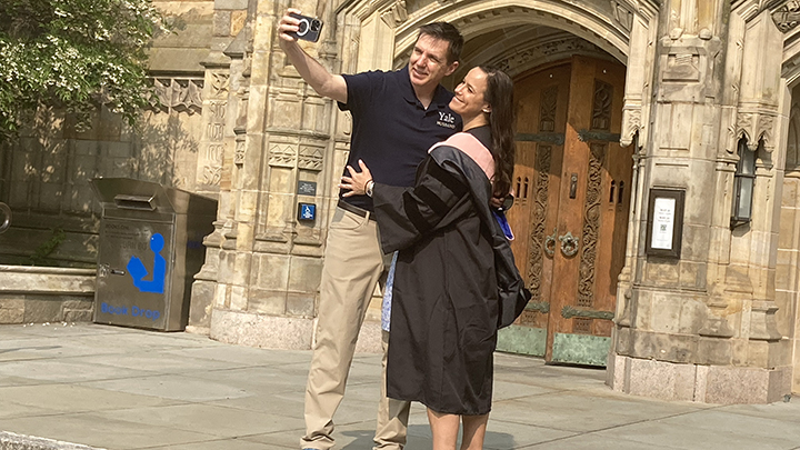 Female graduate with long dark hair hugs man wearing black shirt with Yale written on upper left. He holds a camera and both pose smiling for a selfie