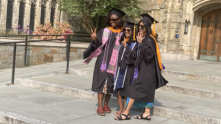 Three women on stone library steps in graduation caps and gowns. Woman at left holds up a peace sign with her fingers, woman at center holds up a single finger, and woman at right waves her full hand at photographer. Woman at far left wears a pink stole with green stripes. All wear sunglasses and sandals.