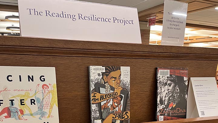 Closeup of book shelves. Top shelf has sign "The Reading Resilience Project." The shelf below has three standing books: The title of the book at center is "I Am Alfonso Jones" showing a graphic illustration of a young man crying, holding an ipad with the image of a black man giving the peace sign.
