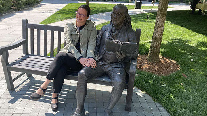 Woman sits in shade on wooden bench, wearing light green jacket and black pants and shoes, hair pulled back, and large dark frame glasses. She has both ands on leg of cast statue of Benjamin Franklin holding book, which sits to her left.