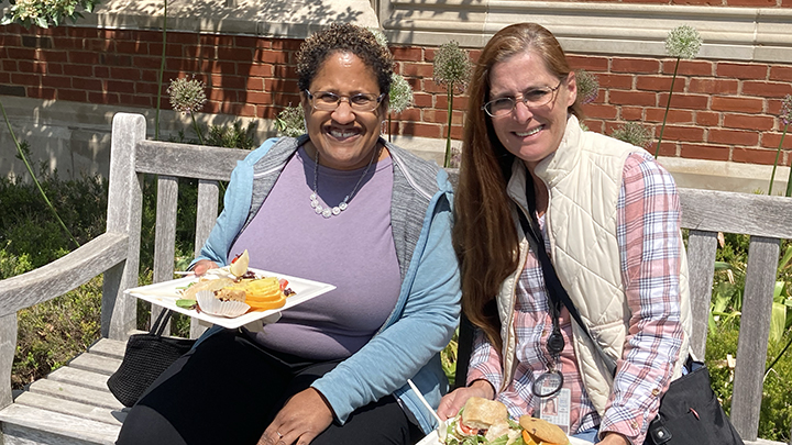 Two women in sunshine on wooden bench. Both wear glasses. Woman at left wears glasses, purple shirt and blue jacket and holds plate of food. Woman at right wears checked shirt and white quilted vest, has long reddish hair and glasses