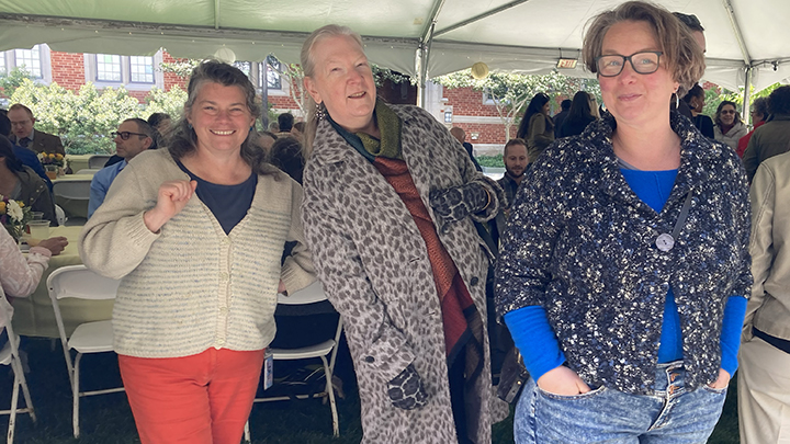 Three woman pose, one at left has white sweater and red pants, woman at center leans toward first and wears animal skin long coat, woman at right has short hair and black-rim glasses and wears a dark cardigan, bright blue top and jeans