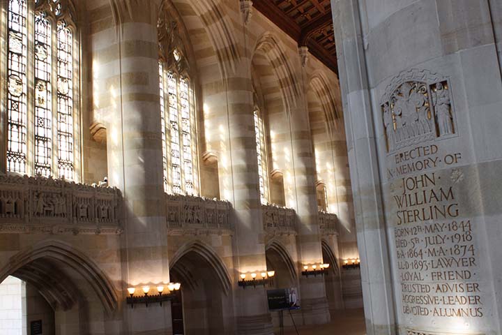 View of soaring two story interior space with stone walls and pillars, painted ceiling,  and stained glass windows. Pillar in foreground has inscription beginning "Erected in Memory of John William Sterling" 