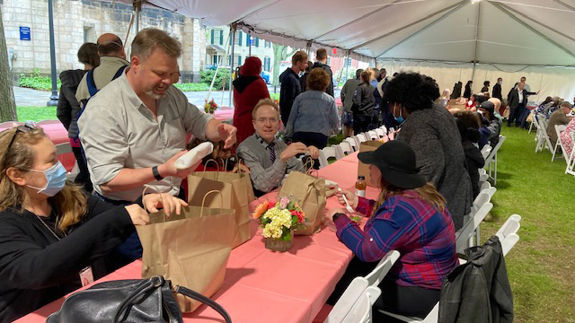 Staff standing and sitting around long pink-covered tables under a tent opening bag lunches. Background shows glimpses of stone buildings and greenery.