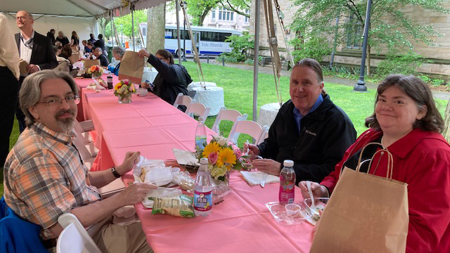 Two men and a woman sitting with bag lunches at long table covered with pink tablecloth under a tent. More staff can be seen in the far background and glimpses of green grass, trees, stone building and a passing bus.