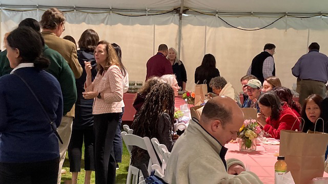 Large group of staff under a tent, some standing and talking, others sitting at long tables with pink tablecloths and flower arrangements. Food and brown paper lunch bags are also on the tables. 