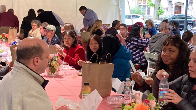 Another view of staff sitting at long pink-covered tables under a tent opening bag lunches. Background shows glimpses of stone buildings and cars on the street.