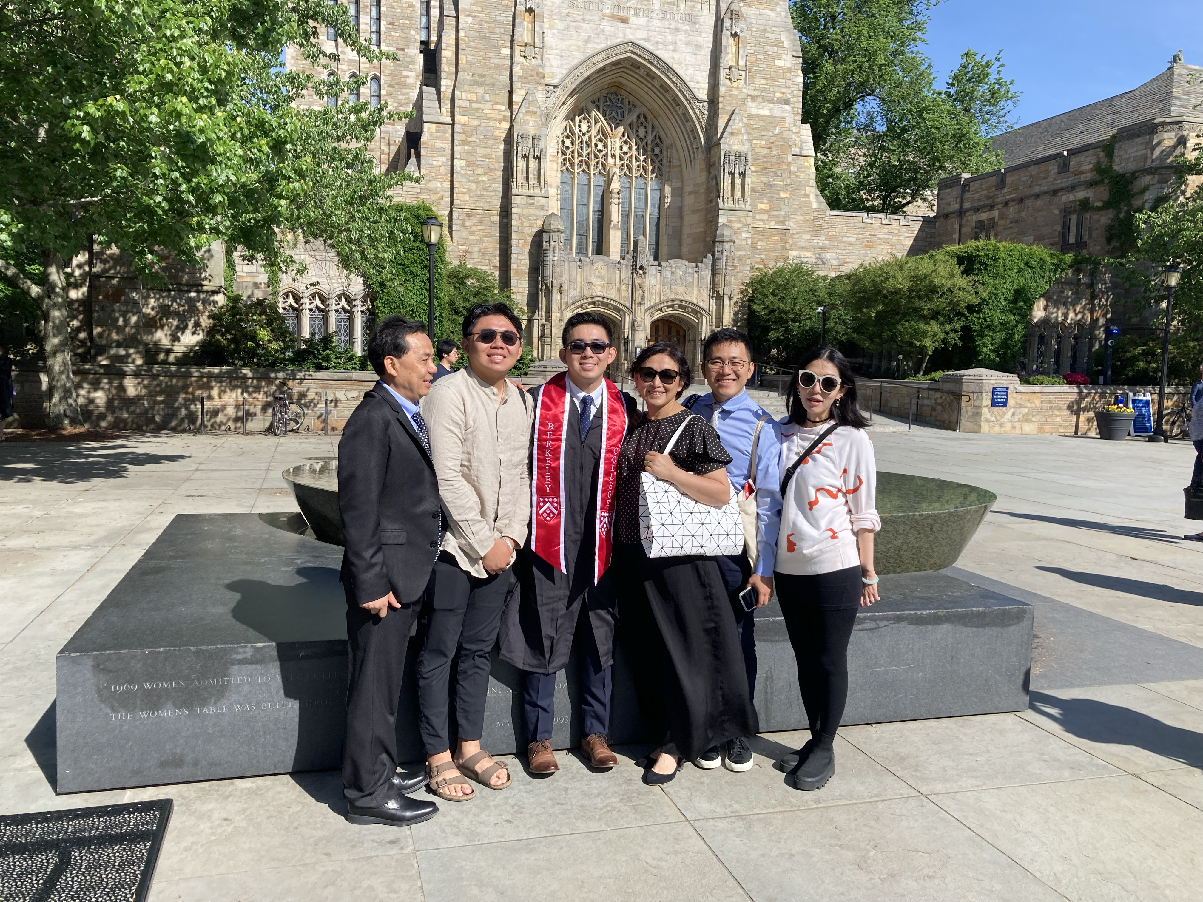 Asian American family of six: The graduate stands at center wearing his gown and the red stole of Berkeley College. To his right are two men, the older one in a dark suit and dress shoes, laughing. The younger one with sandals and a lightweight shirt. The mother wears a black outfit and holds a black and white handbag. Another young man wears a blue shirt. A young girl wears a white sweatshirt with orange design.