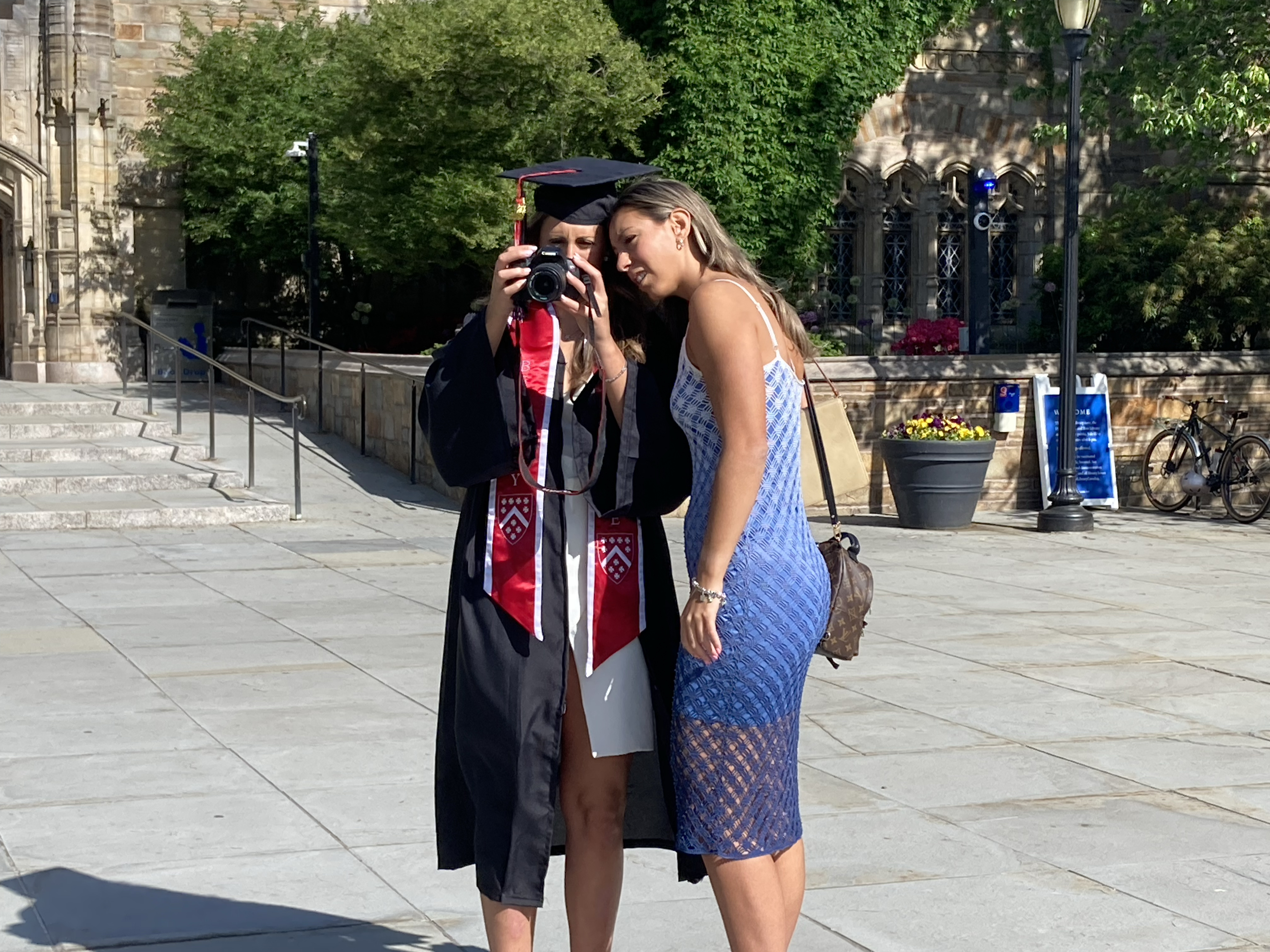 The graduate in cap and gown with red stole and her friend, wearing a blue lacy sundress peer at the back of a camera, heads together, to examine the photo they just took.