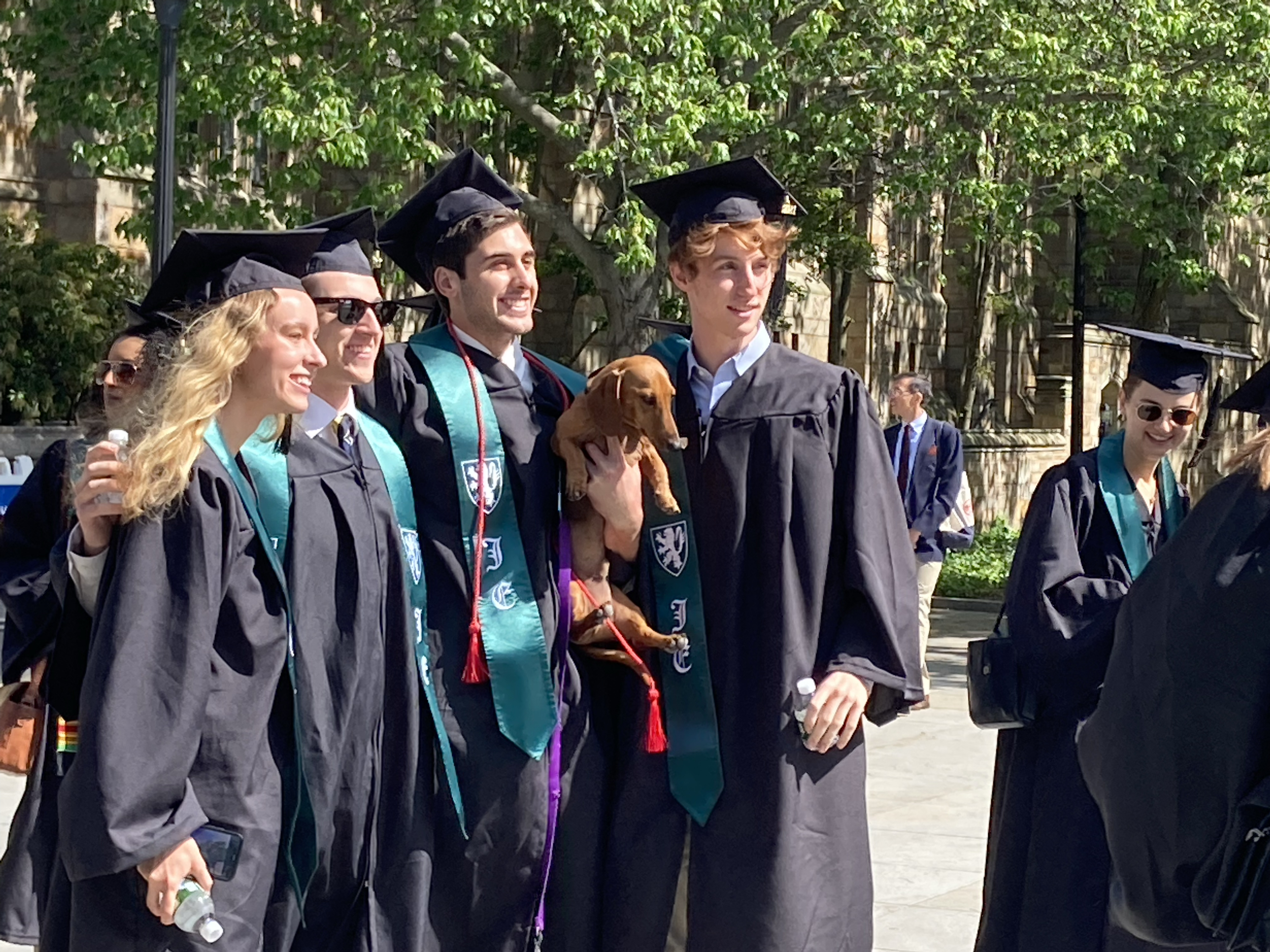Four graduates--a woman with long blonde hair, a man with sunglasses, a man with brown hair holding a brown dachsund puppy, and a man with red hair, all but dog smiling