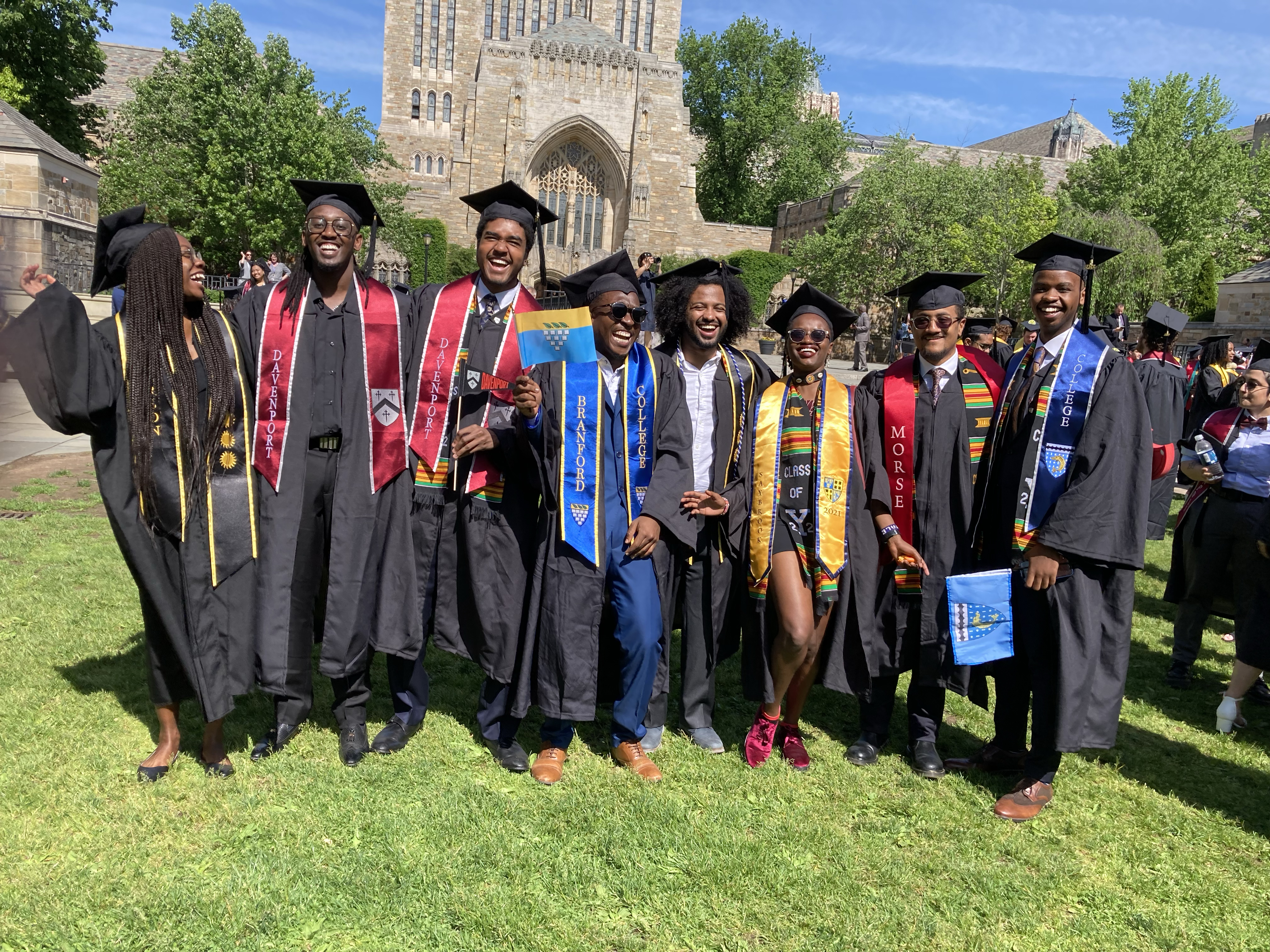 Eight graduates in caps and gowns--from Branford, Davenport, and Morse Colleges--are posing and laughing, several wearing kente cloth stoles.
