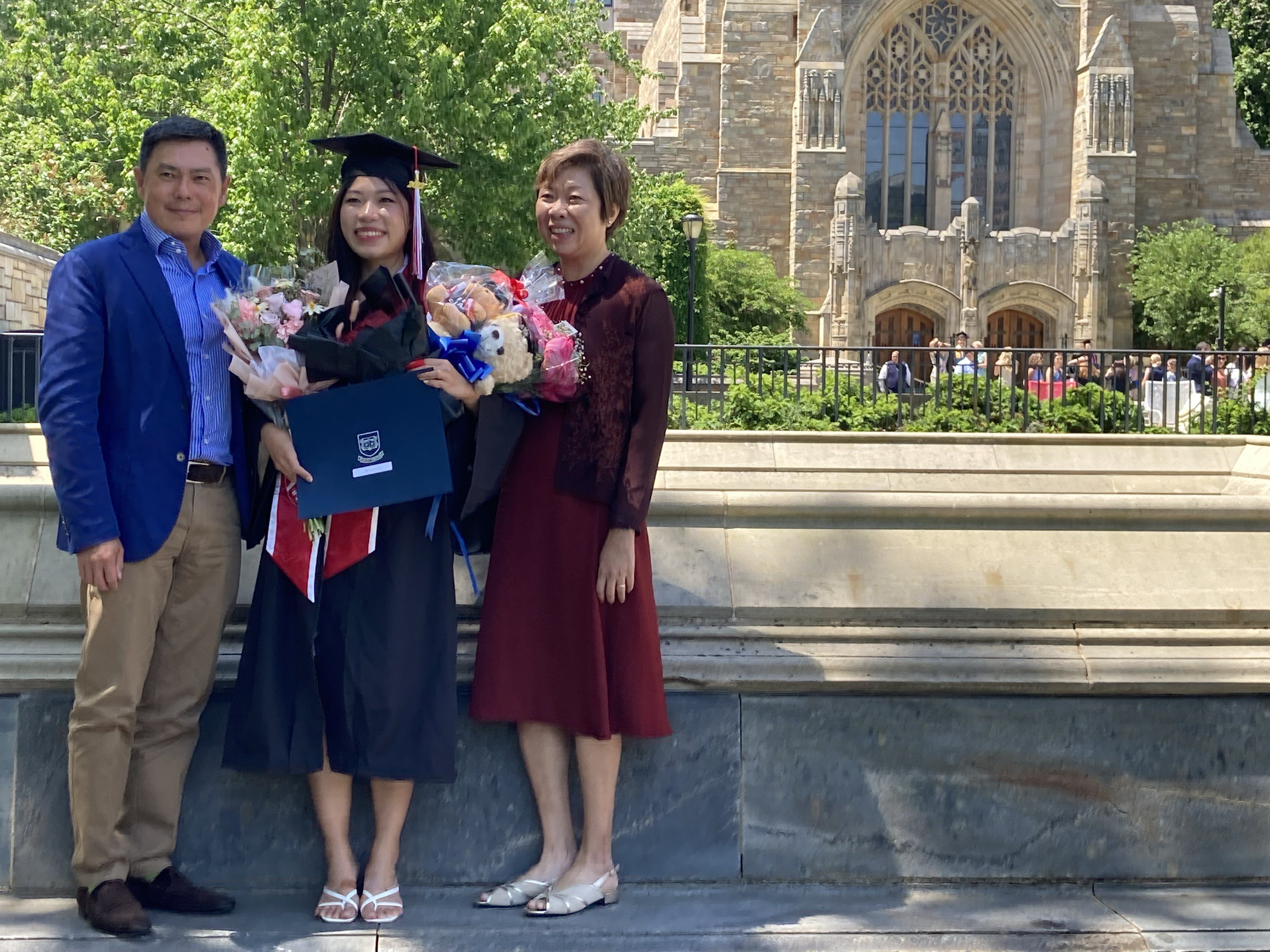 A father, wearing marine blue jacket and blue/white striped shirt, stands next to his graduating daughter. Next to her is her mother, with short brown hair and a burgundy dress and short jacket. The graduate holds three bunches of flowers with bows and teddy bears. She displays the cover of her diploma. The facade of Sterling Library is visible in the background.