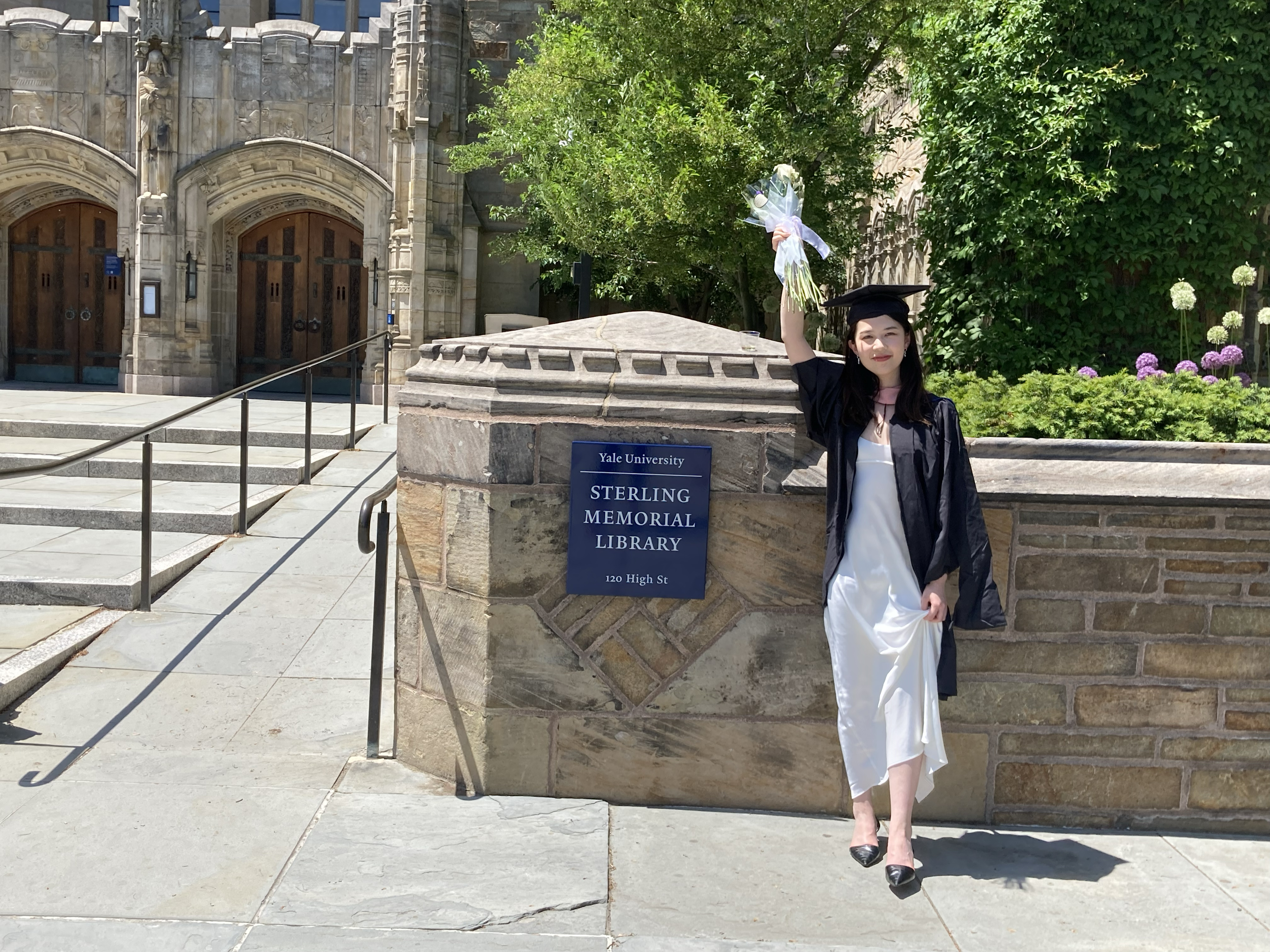 A female graduate wearing a long white dress under her open gown holds a wrapped white rose above her head triumphantly. She stands against a stone wall on which appears the Sterling Memorial Library sign.
