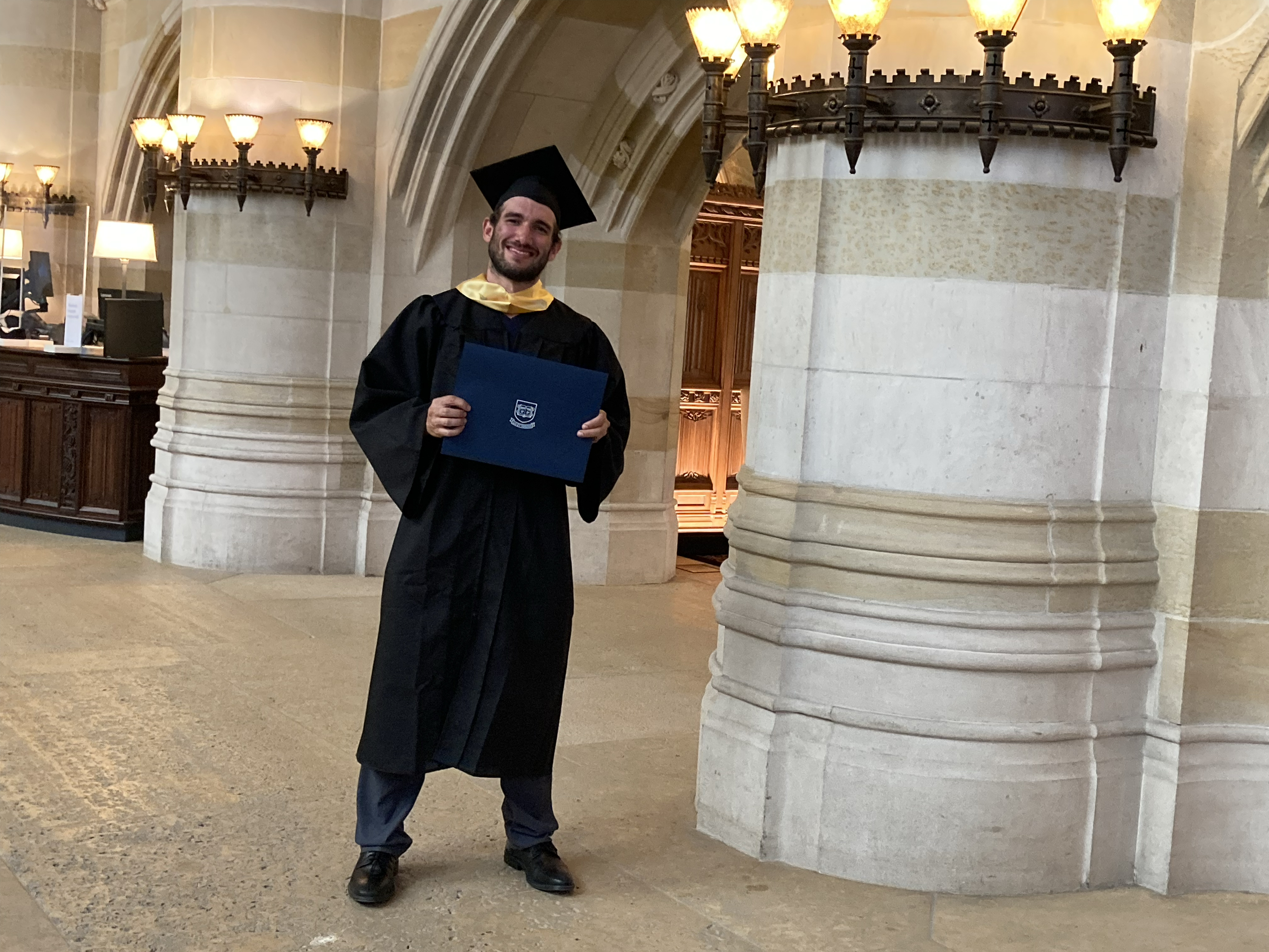 Male graduate wearing yellow stole and holding his blue diploma folder poses in Sterling Nave between pillars near the circulation desk. He has dark hair and a closely trimmed beard and mustache and a large grin.
