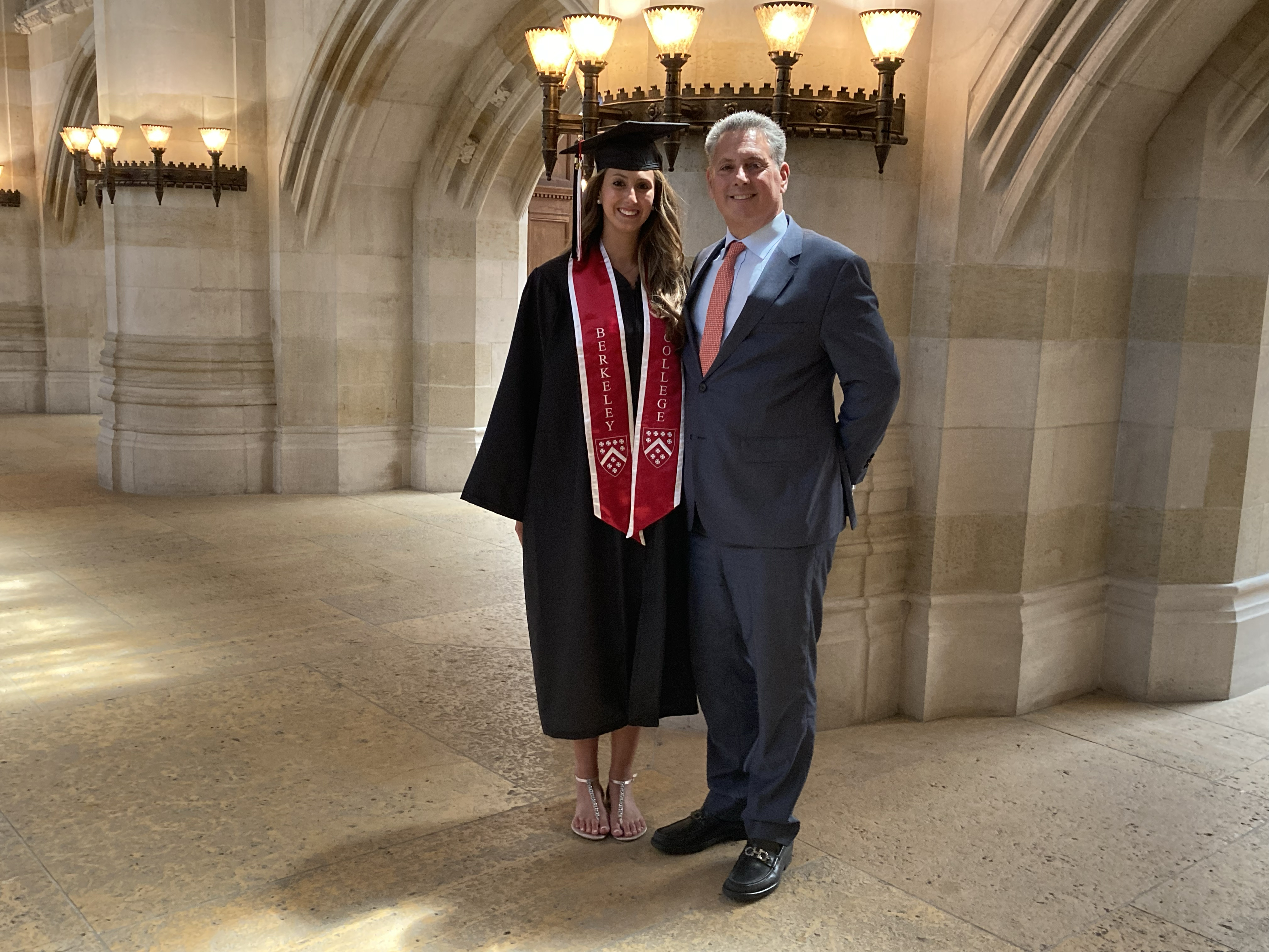 Tall female graduate with long brown hair wearing cap and gown and red Berkeley College stole stands in Sterling nave, next to her proud father, who is wearing a grey suit with blue shirt and red tie. They stand below five lights that curve around one of the nave's columns.