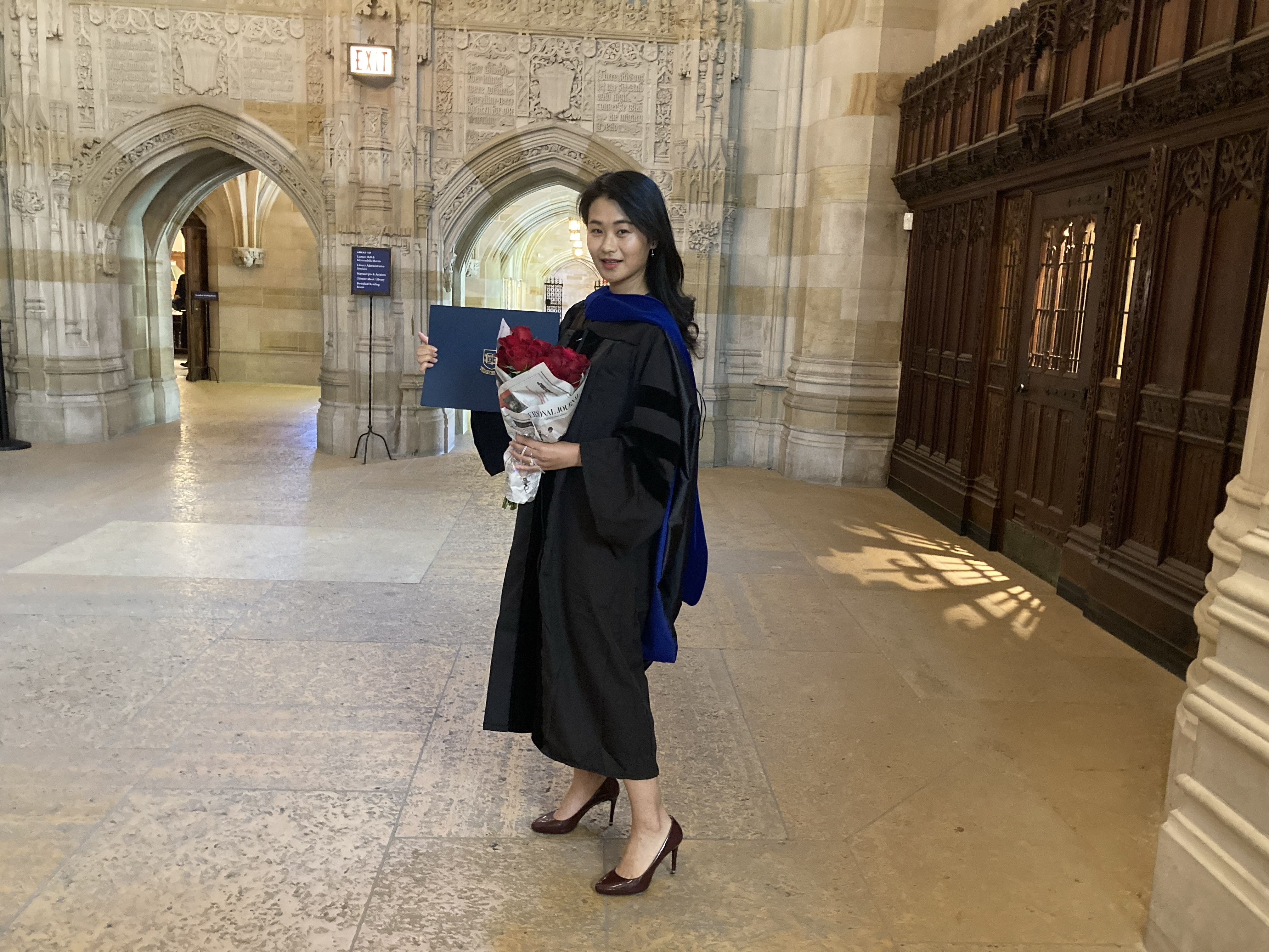 Tall female graduate with long black hair, wearing black high-heel pumps, holds bouquet of red roses wrapped in newspaper and holds her diploma folder. The archways to the exhibition corridor of Sterling Library are visible in the background.