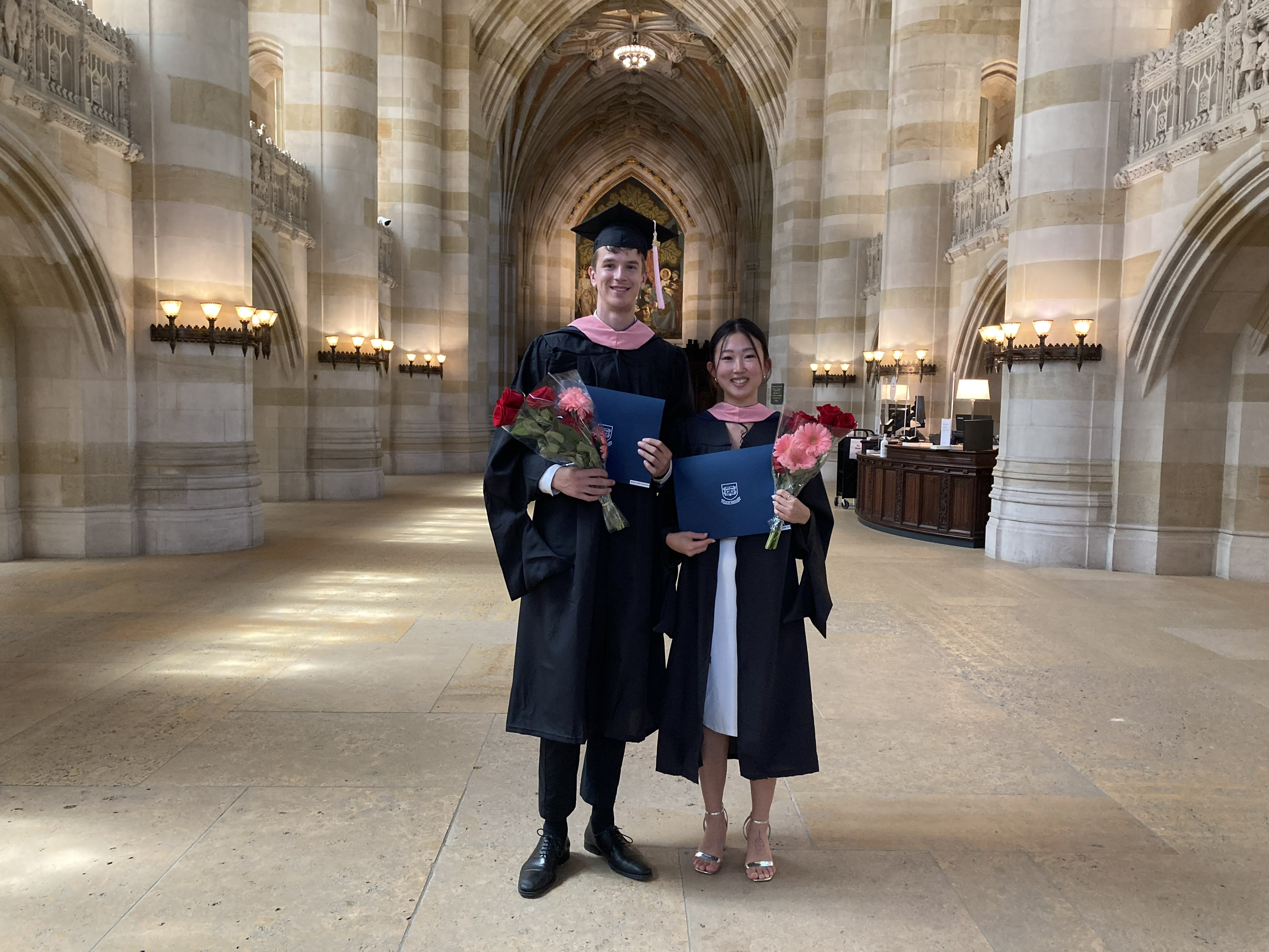 Graduated couple stands in Sterling Nave before painting of Alma Mater. The tall man holds his diploma folder and a bouquet of roses and gerber daisies. The short woman next to him, with black pulled back hair and long side bangs, holds a similar bouquet. Both wear pink stoles and she is wearing a white dress beneath her partially open gown and silver strap sandals.