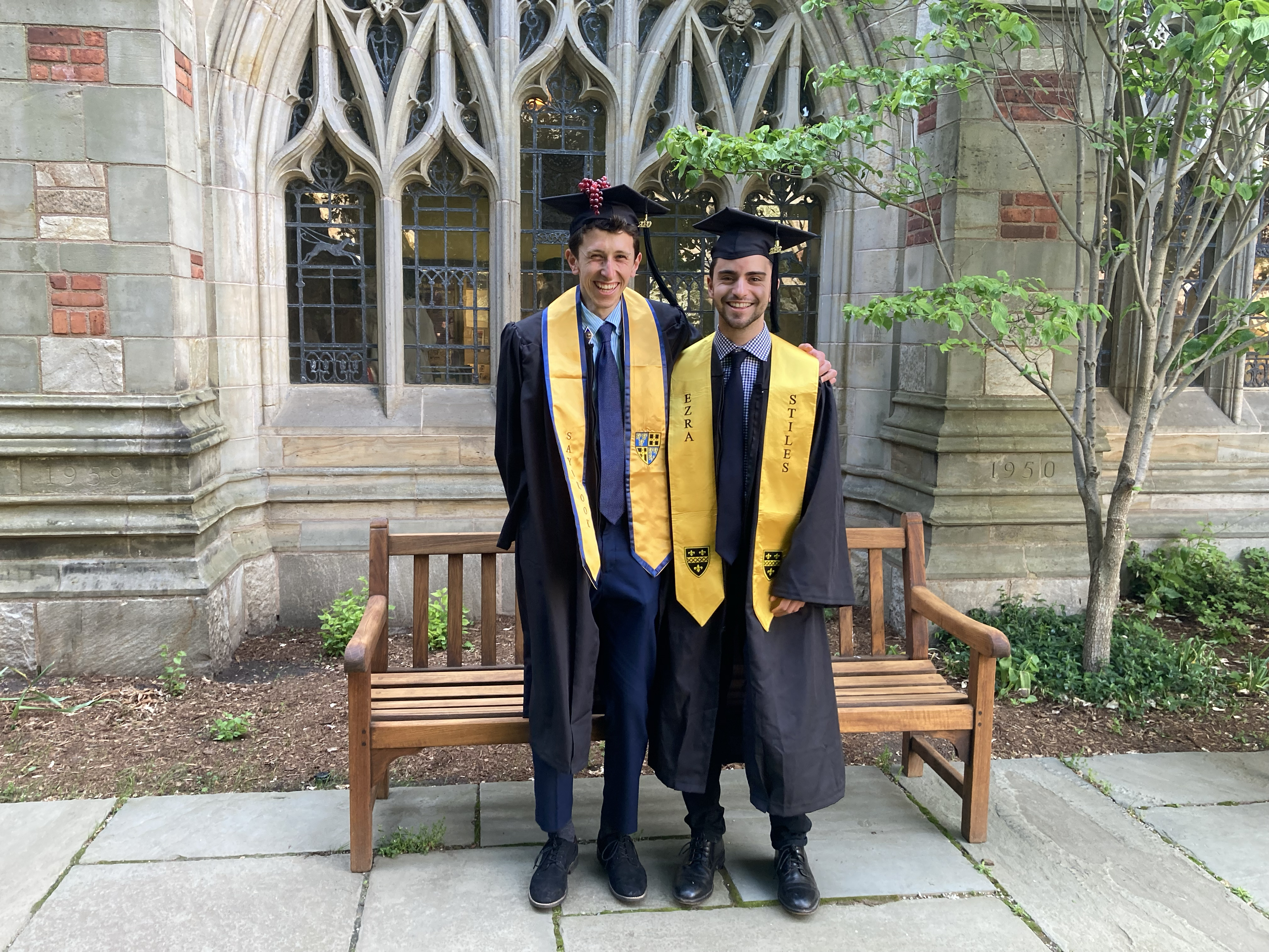 Two friends, both wearing caps and gowns and yellow stoles, pose outside in Sterling's Selin Courtyard. The student at left has grapes attached to his cap and wears the Saybrook College stole. The shorter student at right wears a gold stole for his school and reads "Ezra Stiles." Both men are wearing dark blue ties, visible beneath their partially open gowns. A wooden bench and the leaded glass windows of the Exhibition Corridor are visible behind them. There is a small tree in leaf to the right.