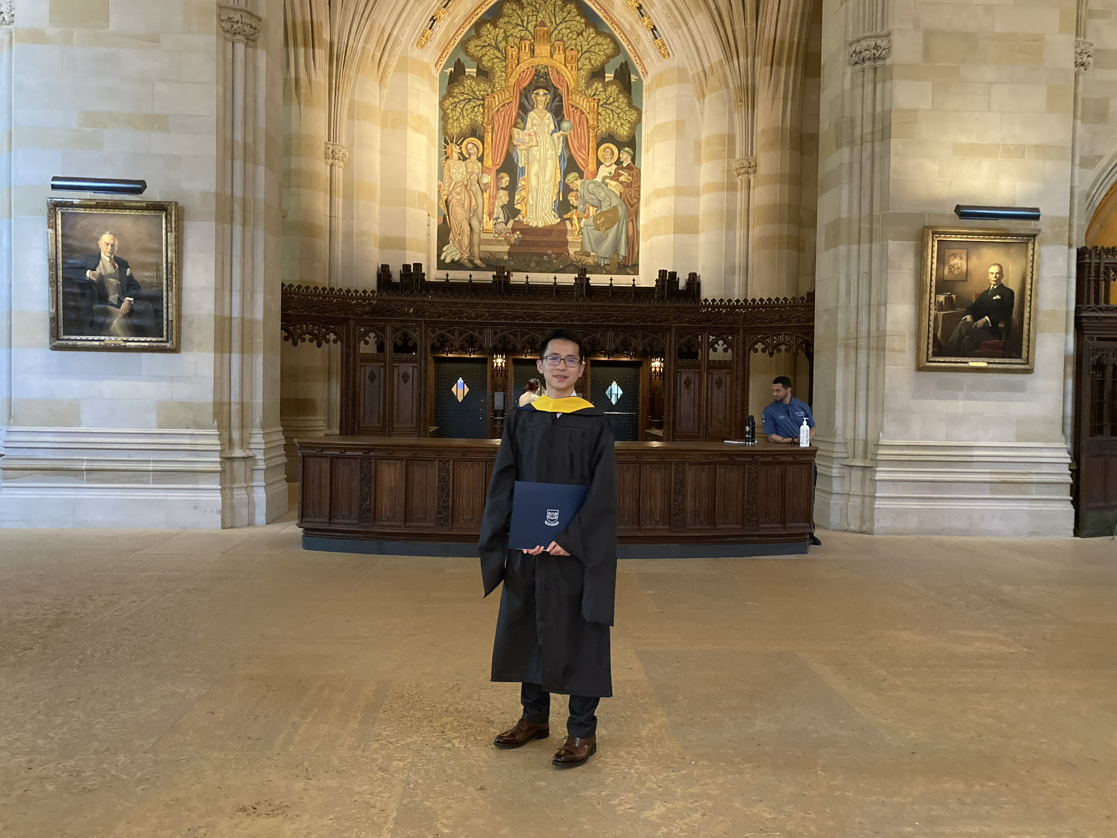 Male graduate without cap, wearing glasses, robe, and yellow stole, holds the blue folder of his diploma, standing in front of the dark wood of the former circulation desk below the painting of Alma Mater in Sterling nave.