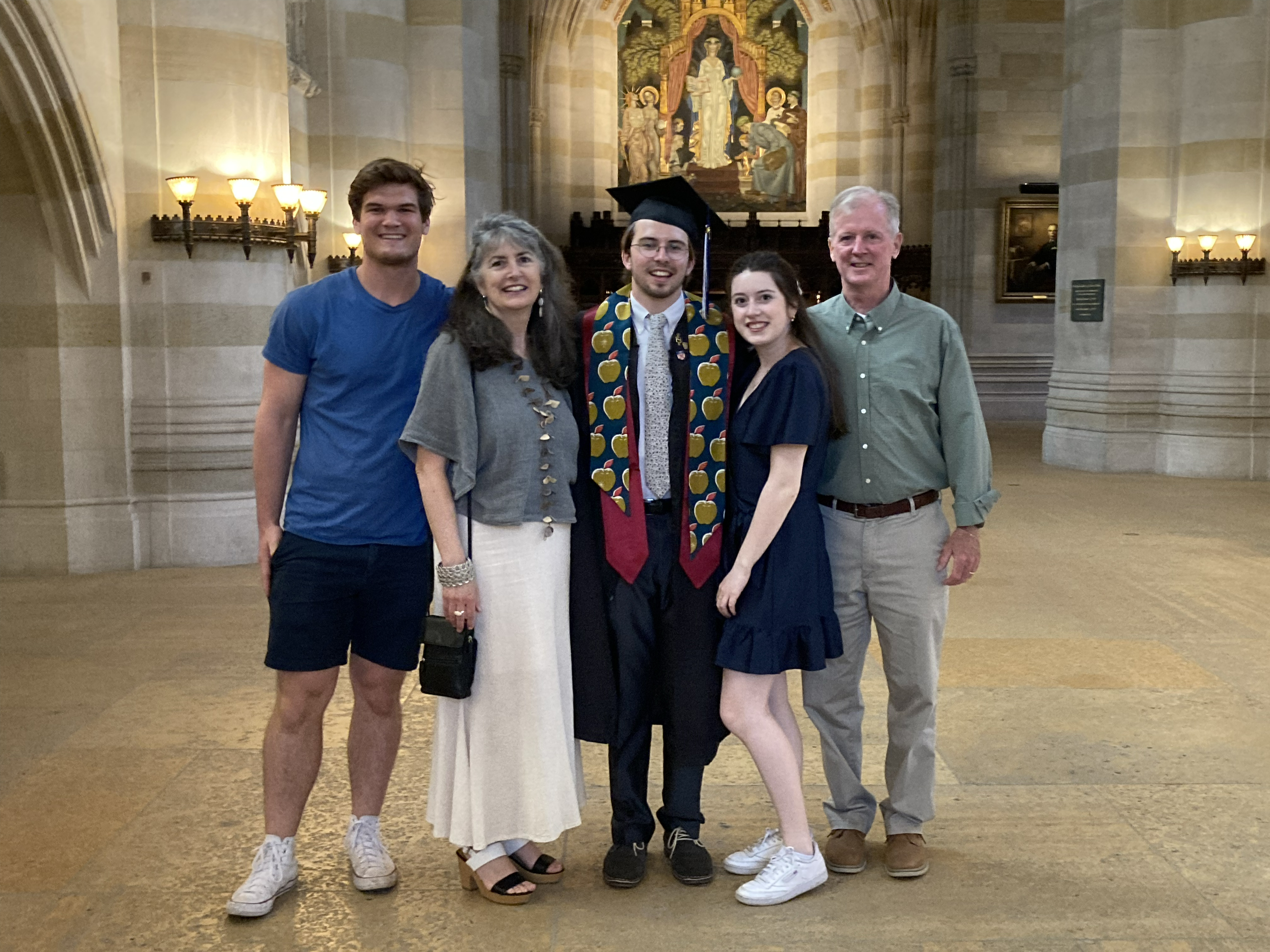 Family of five poses in front of the painting of Alma Mater in Sterling nave: The graduate, at center, wears a short beard and moustache and glasses and a stole embellished with gold apples. A young girl stands to his left, wearing a short blue dress, and an older man in green shirt and khakis stands next to her. To the graduate's right is a woman with long salt-and-pepper hair wearing a gray top and long white skirt; at the end is a tall young man in blue tee shirt and shorts. 