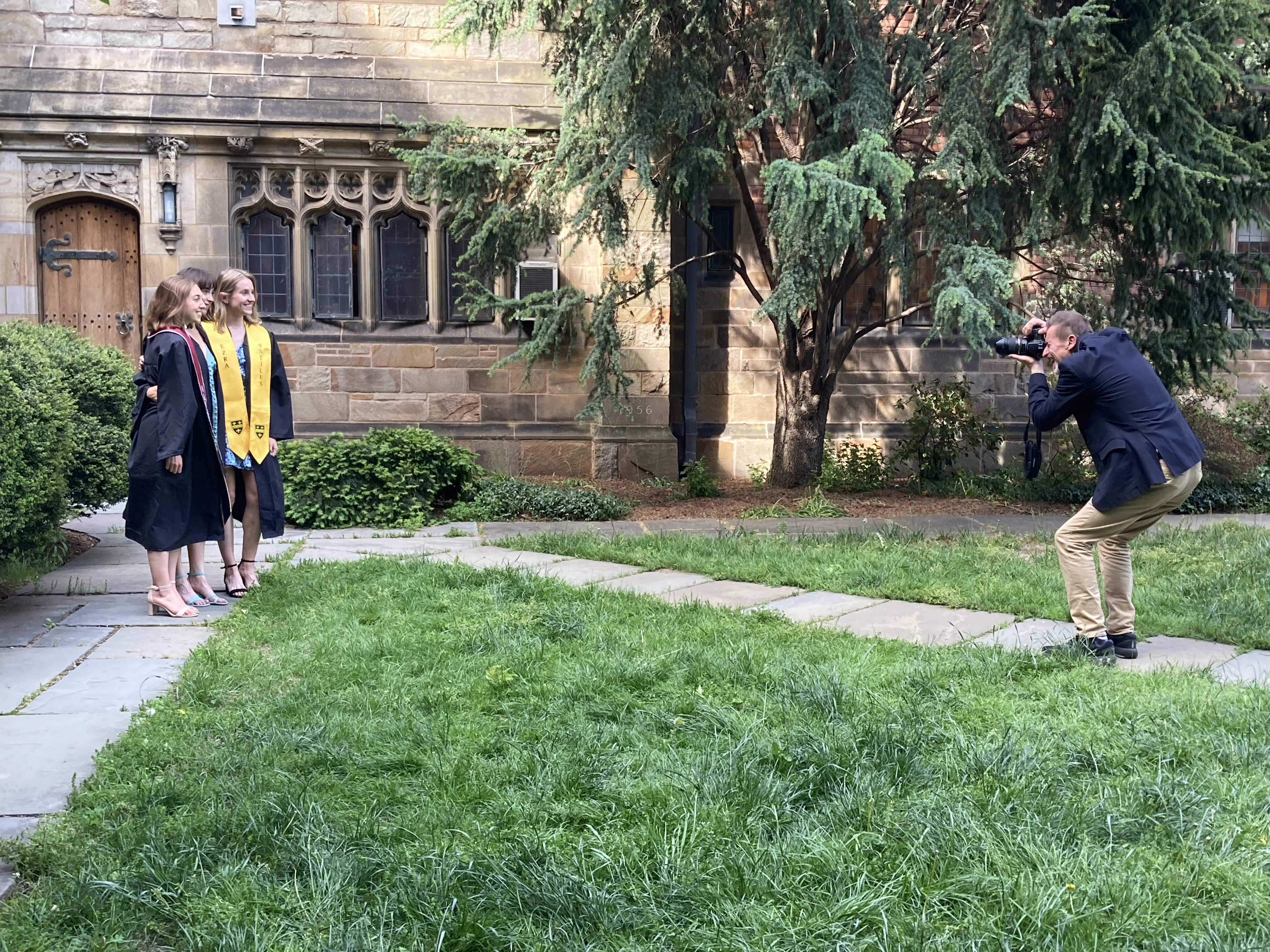A man in dark blue jacket and khaki pants, holding a camera with a long lens to his eye, squats to take a photo of three female graduates posing in Sterling's Selin Courtyard.