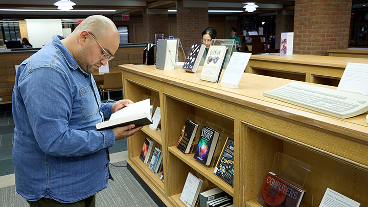 Bald man with glasses and denim shirt stands in front of bookcase reading book with black cover. A keyboard is on the surface of the bookcase along with books on display.