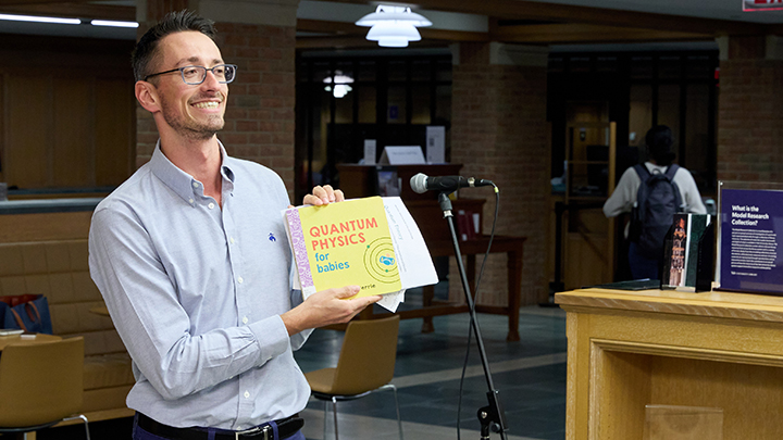 Man with open-neck grey shirt and glasses holds up yellow book that reads "Quantum Physics for Babies"