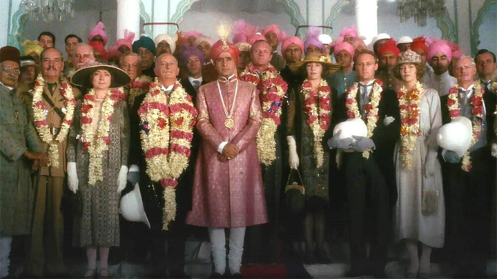 Photo of large group facing camera, wearing early 20th century clothing. Many wear red and white flowered leis. Man at center is in pink nehru jacket with red turban with feather and gold medallion on long white necklace.