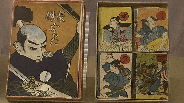Large painted card on left shows bald Japanese warrior wielding sword. At right four smaller playing cards each show a Japanese man in traditional dress. Each card has a red circle with a Japanese character at center.