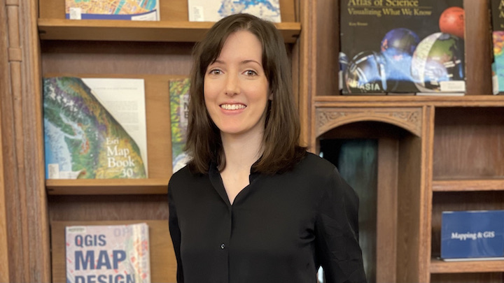 Young woman with shoulder length dark hair wearing black collared shirt stands in front of book shelves showing books with covers facing out. 