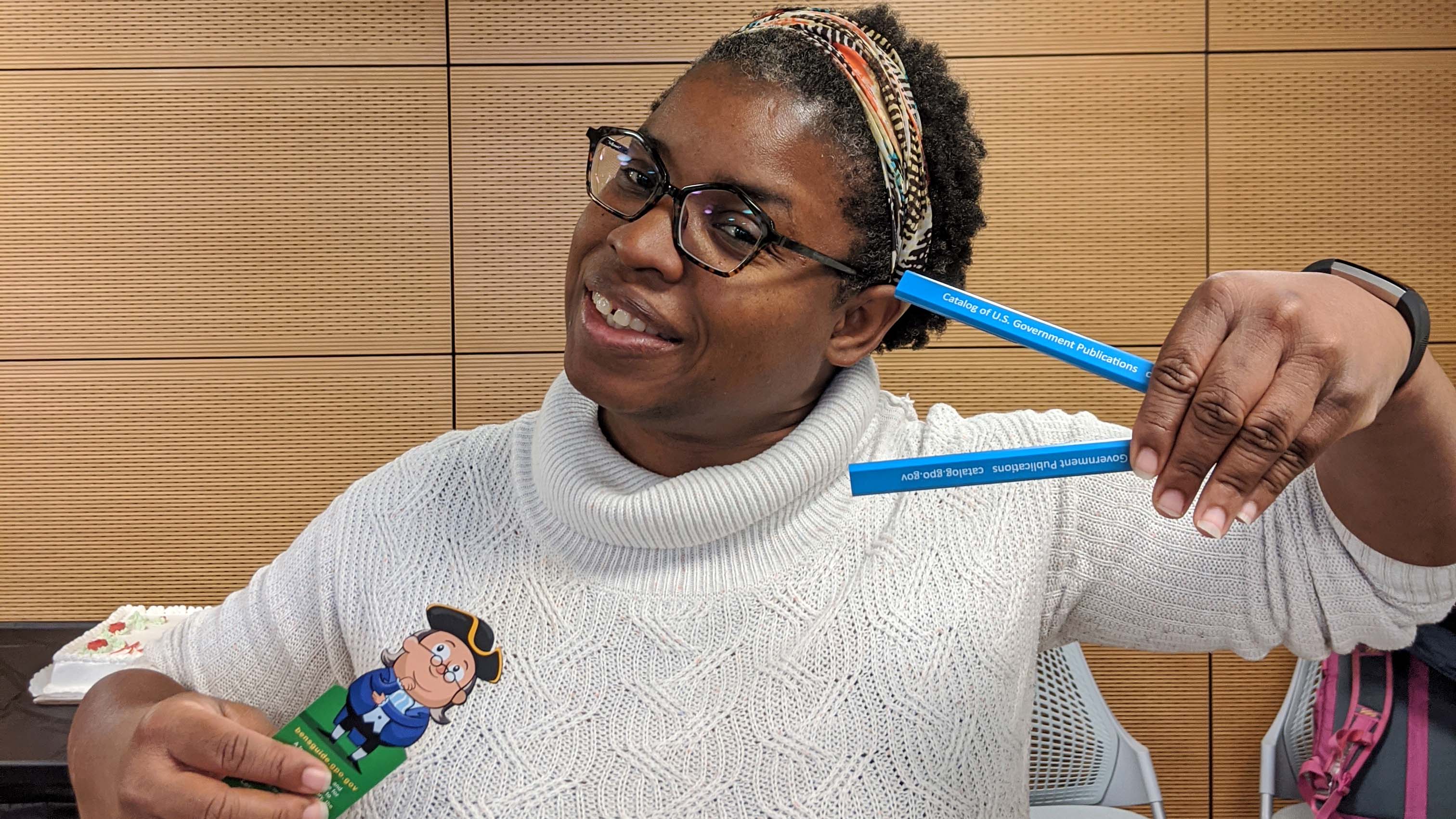Smiling Black woman wearng white turtleneck sweater, glasses, and colorful hairband, holding pencils in one hand and a bookmark in the other.