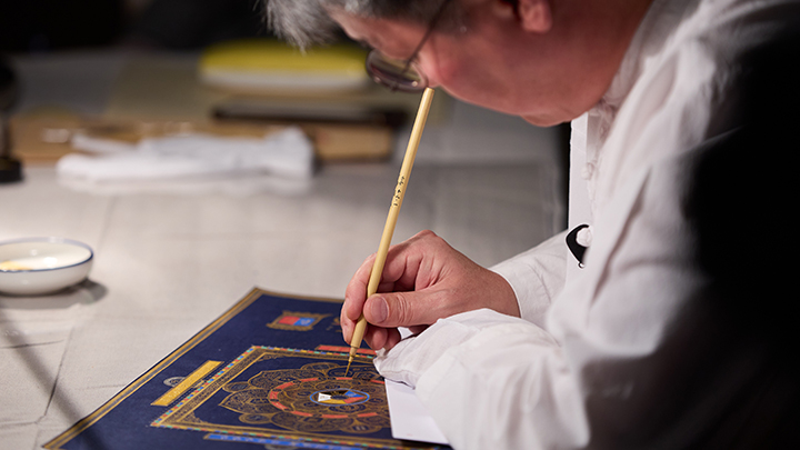 Man bends over red, blue and gold illuminated drawing holding bamboo pen upright in right hand