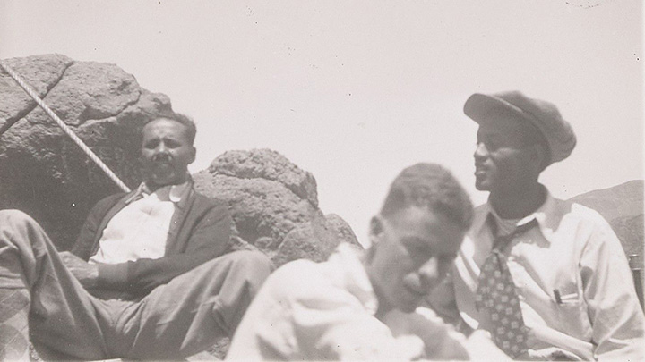 three men  resting against rocks in black and white photo. One at left looks at camera and wears dark cardigan; man at center looks down, and man at right wears cap and wide patterned tie with pen in his shirt pocket