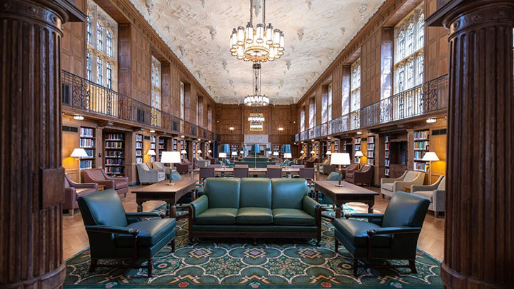 Wide view of room with green couches in the foreground, three round chandeliers and wood paneling and bookshelves on either side