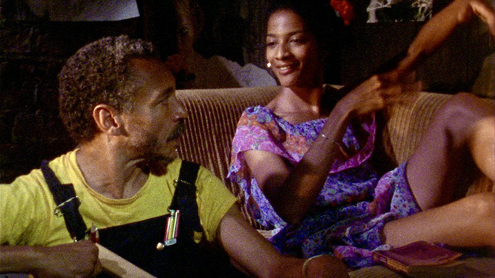 Man in black overalls and yellow t shirt looks at a woman in a purple print dress sitting next to him.