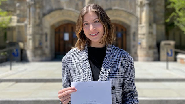 Female student stands in front of Sterling Memorial Library. She has chin-length brown hair parted in the center, and wears a black collarless shirt under a black-and-white plaid blazer. She holds a copy of her paper in front of her.